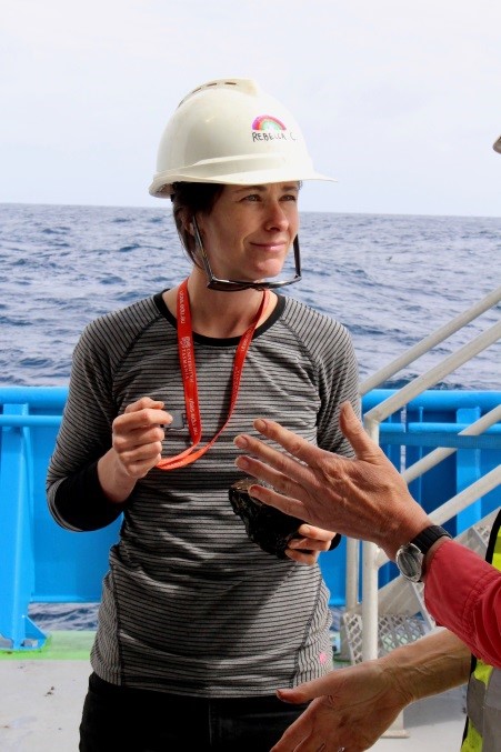 woman stands on ship deck in hardhat and striped long sleve t-shirt with sea behind her