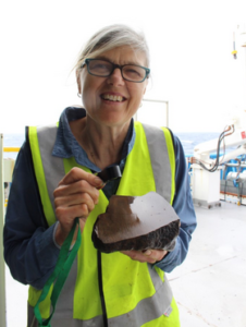 woman in yellow high vis vest and glasses holds rock and brushing tool and smiles at camera