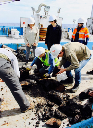 six scientists in hard hats look through a pile of rocks on the ship deck