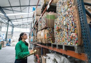 woman with clipboard looks up at pallets of packed plastics on a shelf