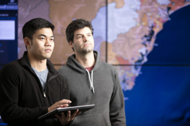 Two young men, one with a tablet, standing in front of a map displayed on multiple screens.